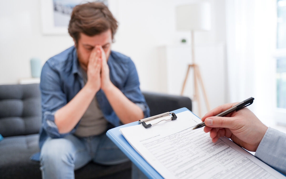 Man with head in hands showing signs of low testosterone and mental health distress while a provider takes notes during a clinical consultation